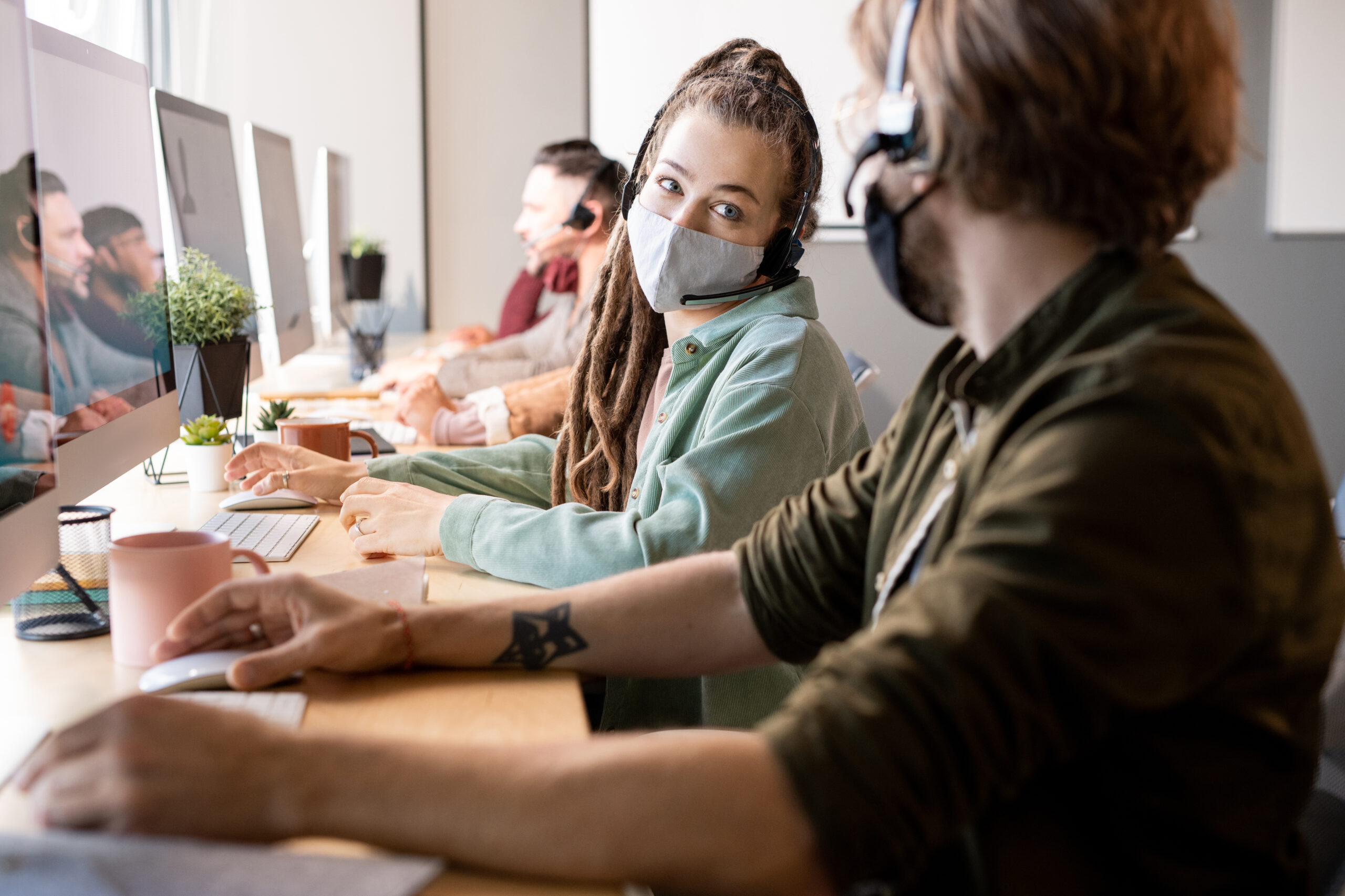 Young female operator of call center in protective mask looking at one of colleagues while consulting with him about client question